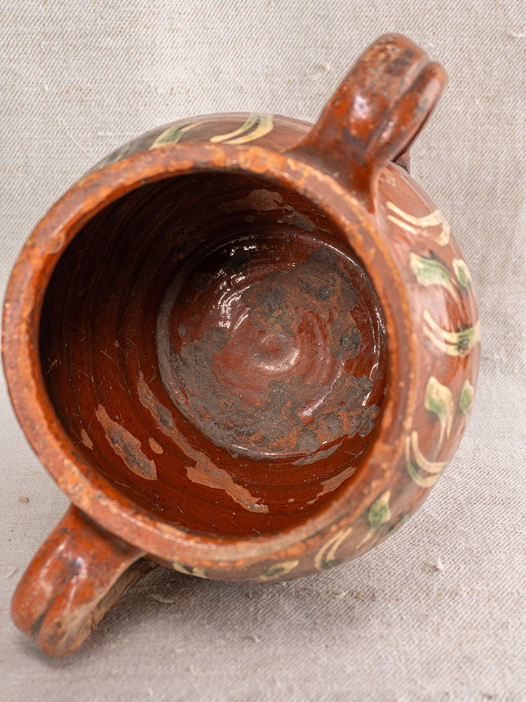 Early 19th century French terracotta slipware pot with two handles, glossy interior, and green-yellow patterned rim on beige fabric.