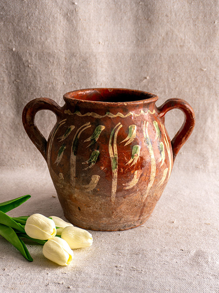 French terracotta slipware pot with jaspé green-white patterns and two handles, displayed on beige fabric with white tulips.