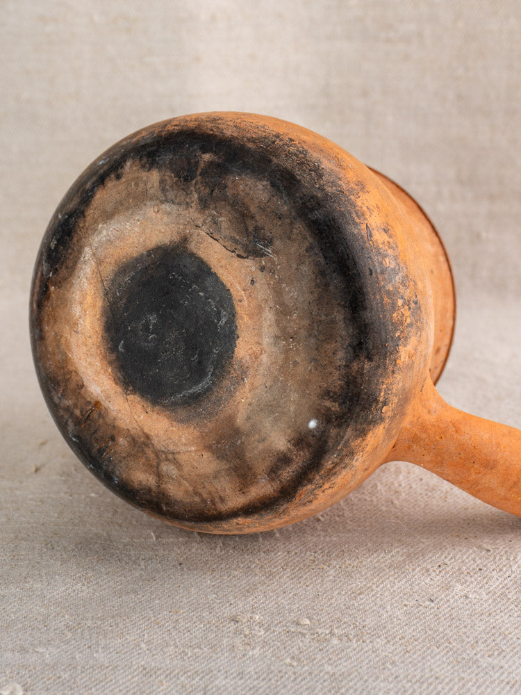 Bottom of antique French Vallauris terracotta poêlon pot with hollow handle, burn marks, on neutral fabric.