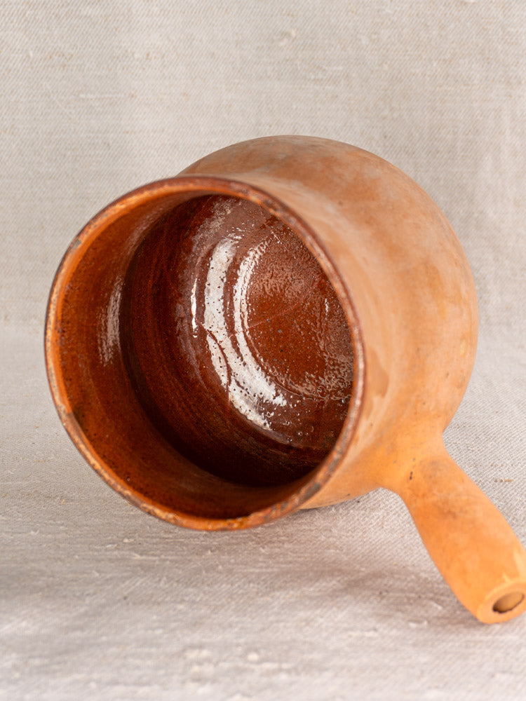 Antique French Vallauris terracotta poêlon with hollow handle, glossy interior displayed on neutral fabric background.