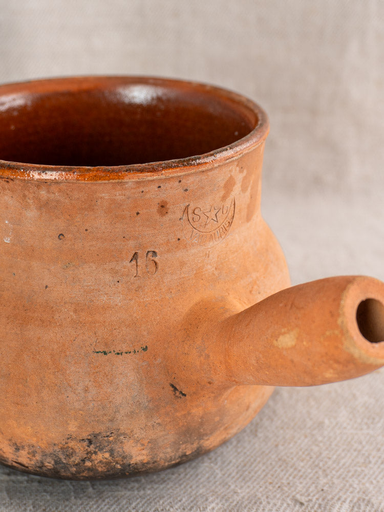 Antique French Vallauris terracotta poêlon with hollow handle, “16” stamp, and glazed interior on textured fabric background.