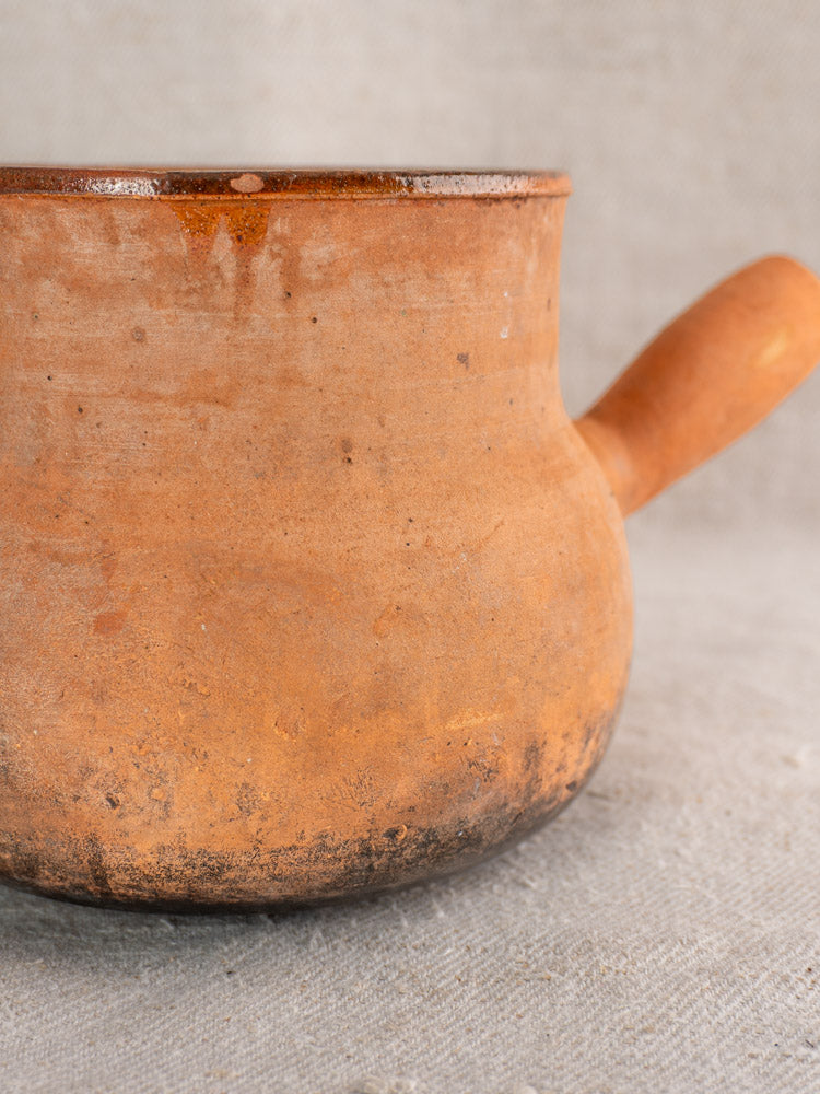 Antique French Vallauris terracotta poêlon with hollow handle, weathered clay and darkened base, set on light fabric.