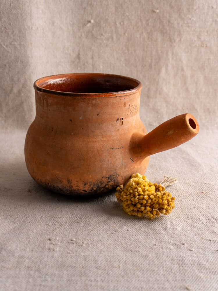 Antique French Vallauris terracotta poêlon cooking pot with hollow handle on beige fabric beside dried yellow flowers.