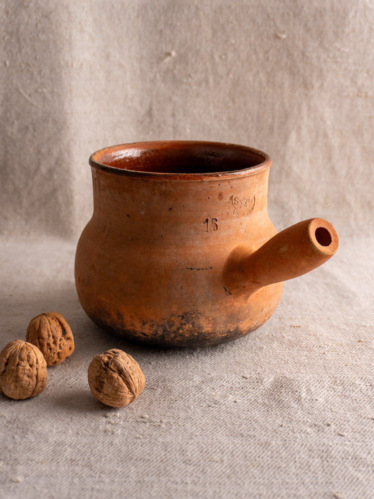 Antique French Vallauris terracotta poêlon with spout and hollow handle on textured fabric, walnuts beside.
