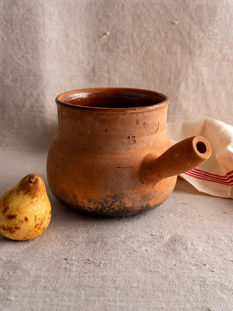 Antique French Vallauris terracotta poêlon pot with hollow handle on fabric beside pear and folded white cloth.