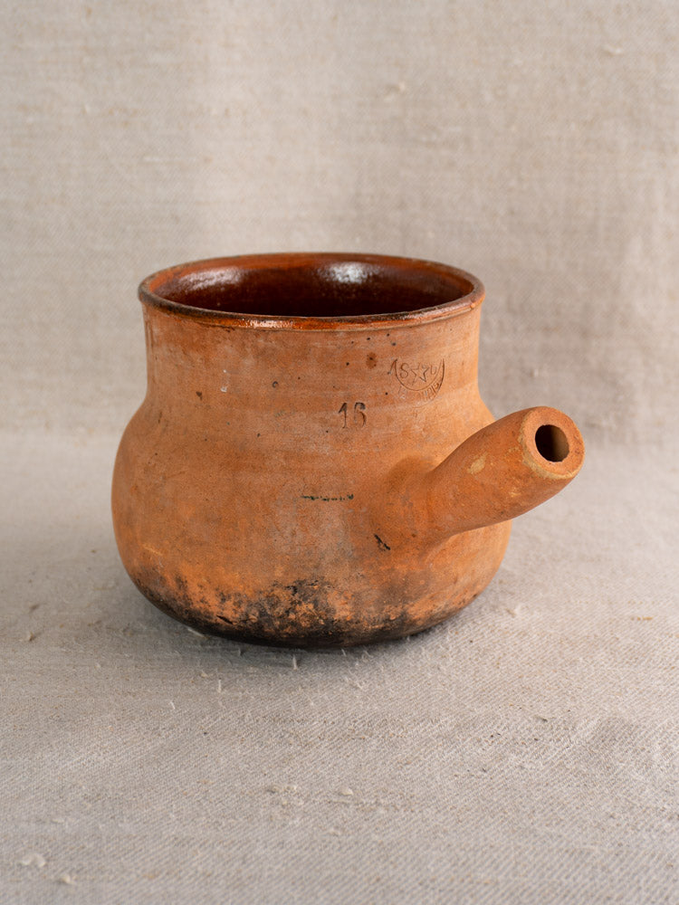Antique French Vallauris terracotta poêlon cooking pot with hollow handle on textured beige fabric background.
