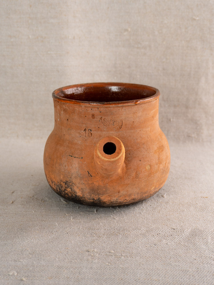 Antique French Vallauris poêlon terracotta cooking pot with hollow handle, short spout, and reddish-brown patina on beige fabric.