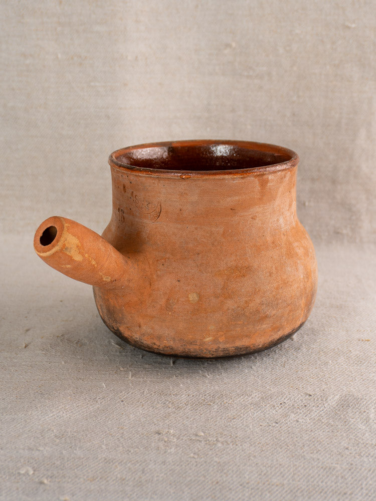 Antique French Vallauris Poêlon terracotta pot with hollow handle on beige textured fabric, rustic earthy brown finish.