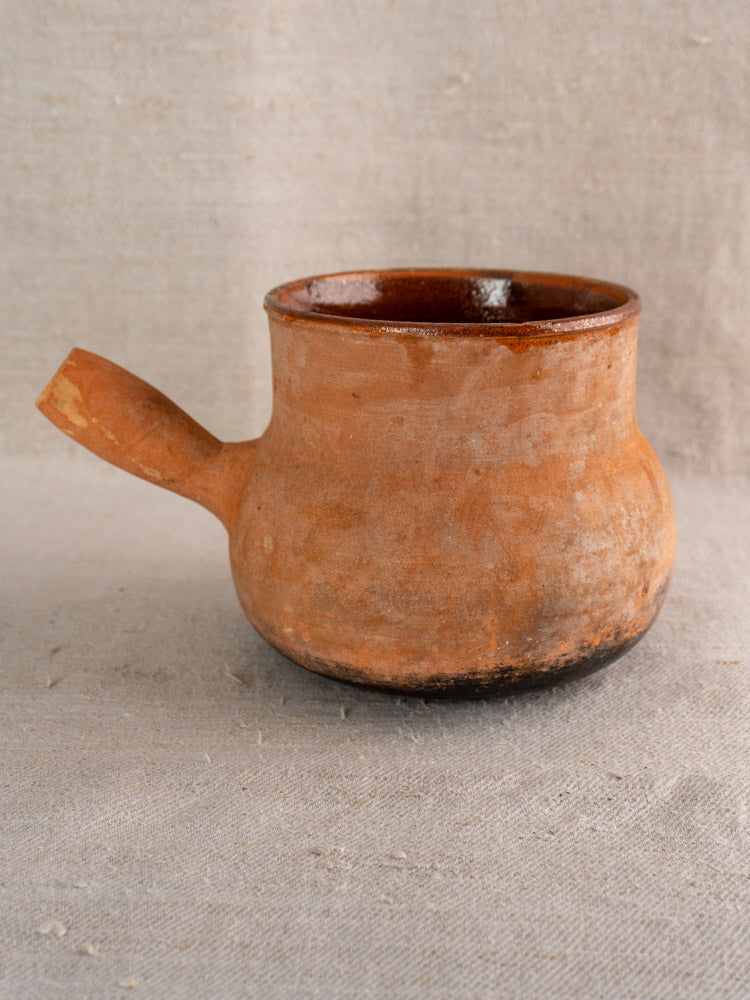 Antique French Vallauris terracotta poêlon with hollow handle and rustic finish on textured light gray fabric.