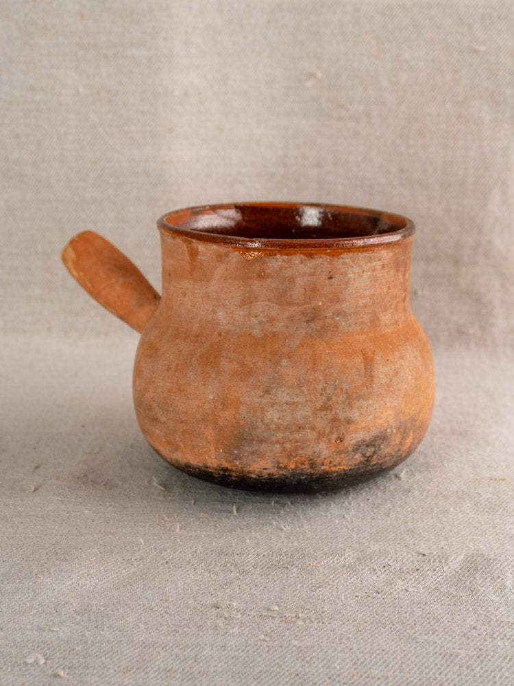 Antique French Vallauris terracotta poêlon with hollow handle and rounded body on textured beige fabric, rustic aged finish.
