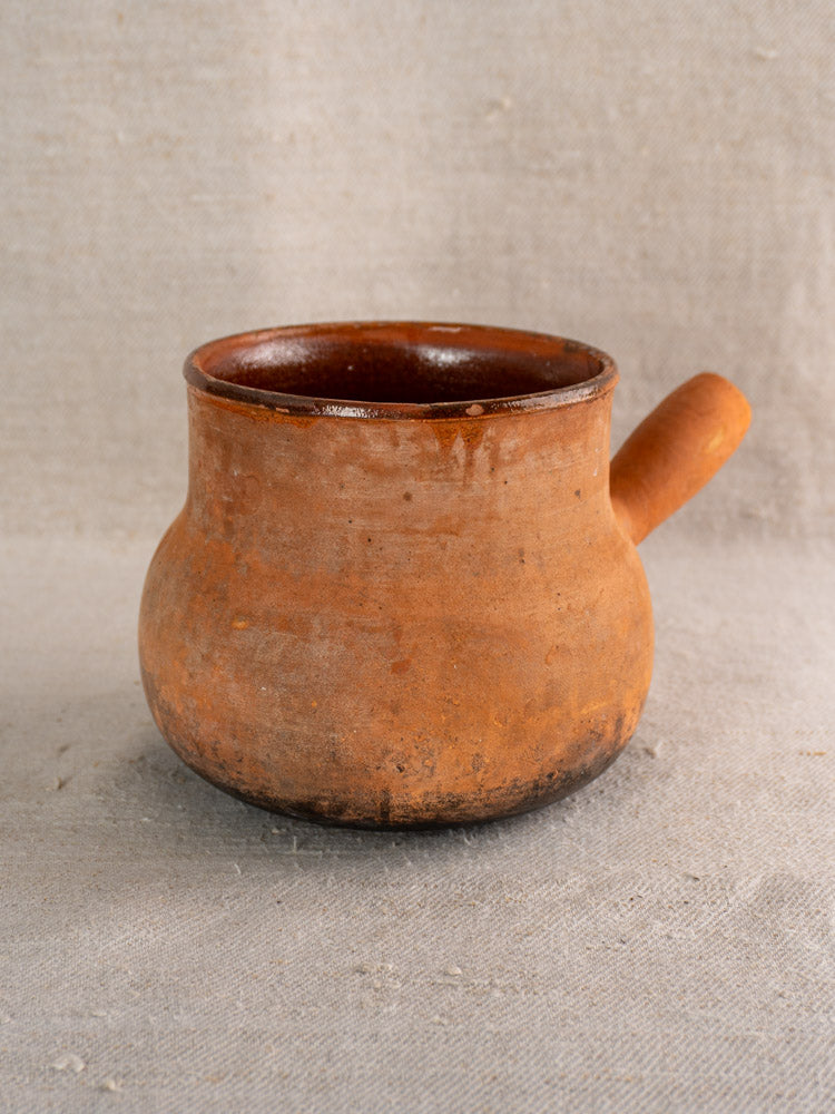 Antique French Vallauris terracotta poêlon with hollow handle and wide opening on textured beige fabric.