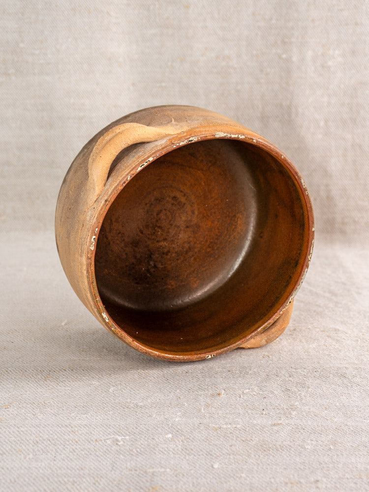 Handmade brown ceramic bowl with two handles, fire-darkened glaze, on textured beige fabric.