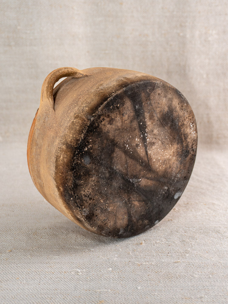 Antique French glazed terracotta hearth cooking pot with fire-darkened base on neutral fabric background.