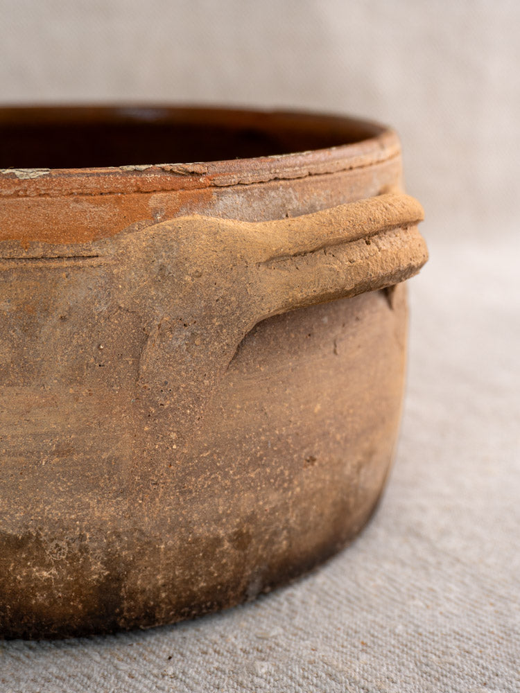 Antique French terracotta hearth cooking pot with rustic brown glaze and aged, textured surface on woven fabric.