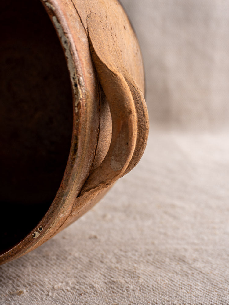 Antique French terracotta cooking pot with curved handle, rustic fire-darkened glaze on beige fabric.