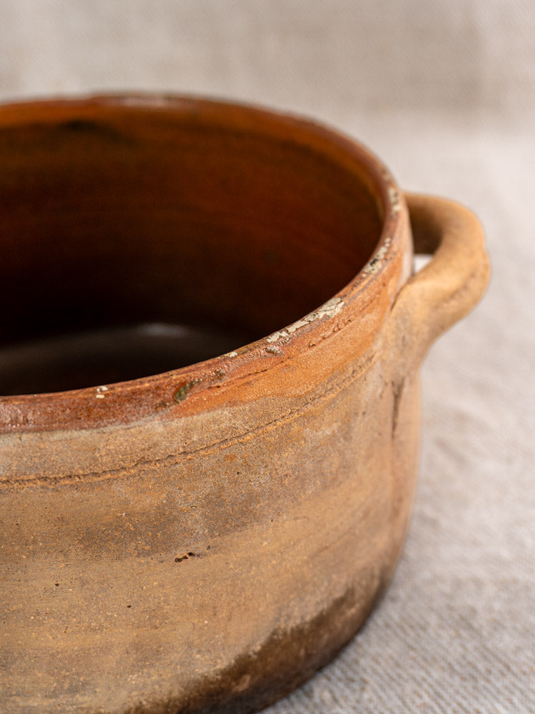Antique French terracotta cooking pot with rustic handle and fire-darkened glaze on fabric background.