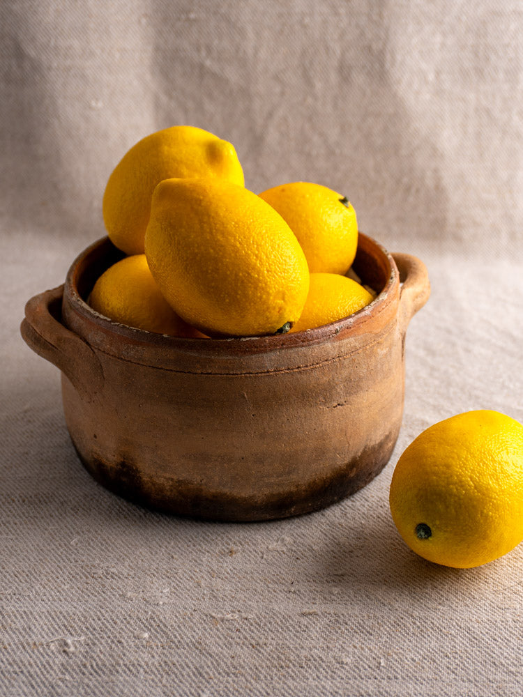 Antique French terracotta hearth cooking pot with fire-darkened glaze, filled with lemons on beige fabric.