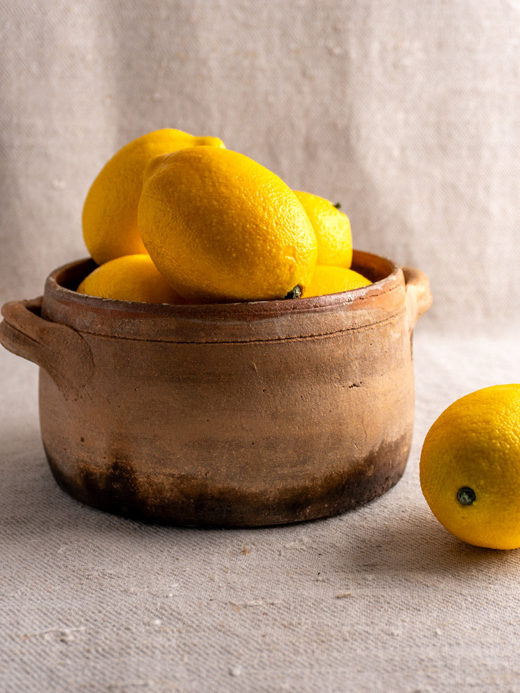 Antique French terracotta hearth cooking pot in rustic brown, filled with lemons on beige textured fabric.