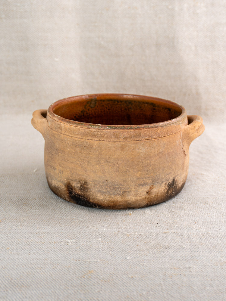 Antique French terracotta hearth cooking pot with side handles and fire-darkened base on textured surface.