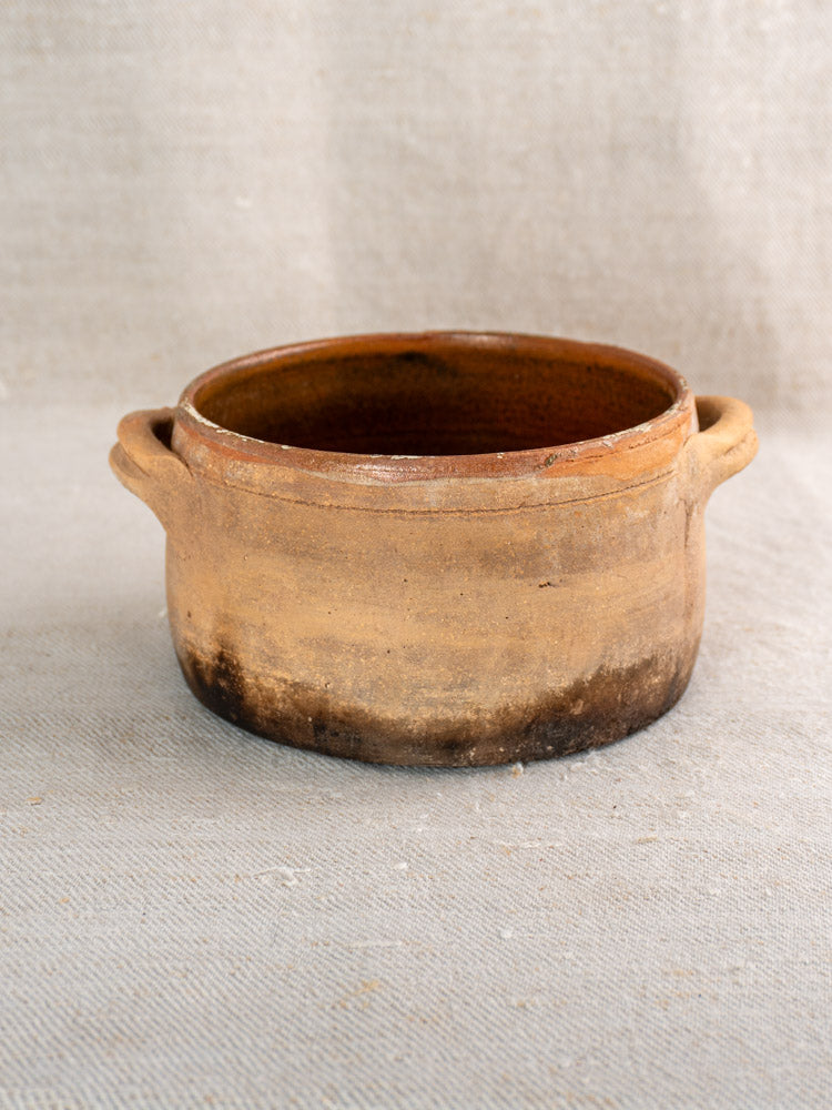 Antique French terracotta hearth cooking pot with brown glaze and fire-darkened base, on neutral fabric.