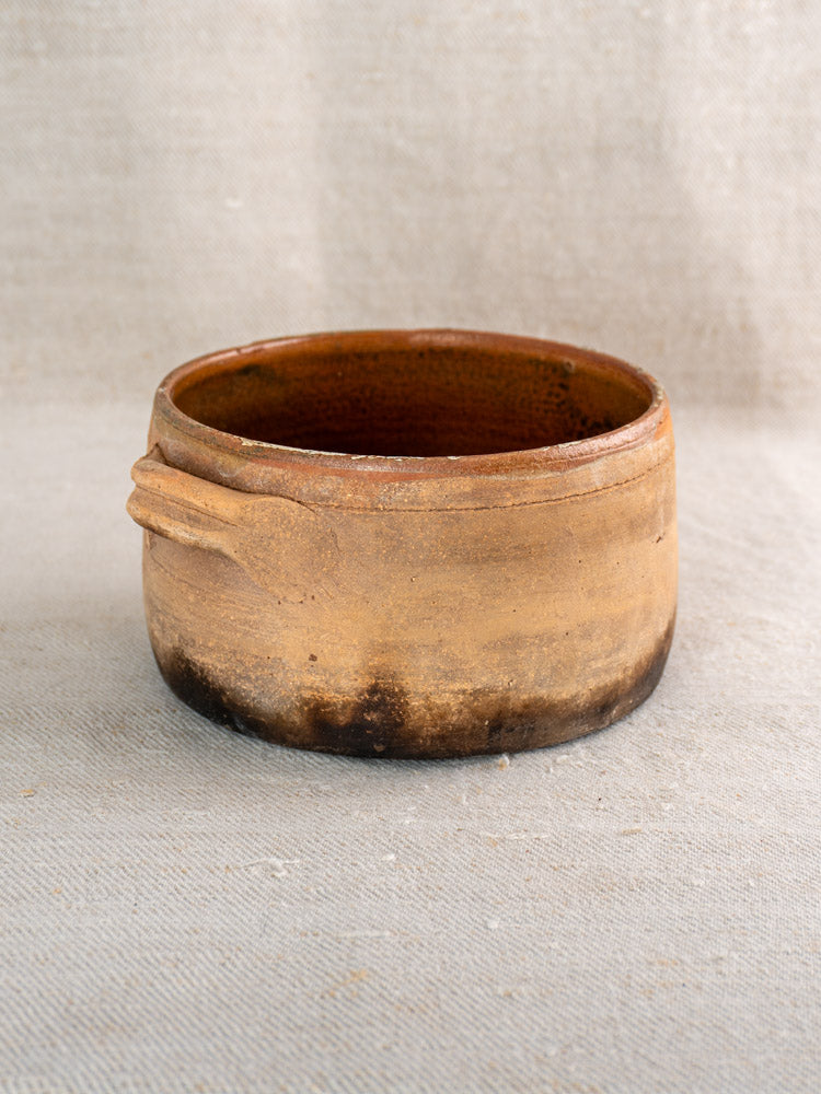 Antique French terracotta hearth cooking pot with glazed, textured surface and side handle on neutral fabric.