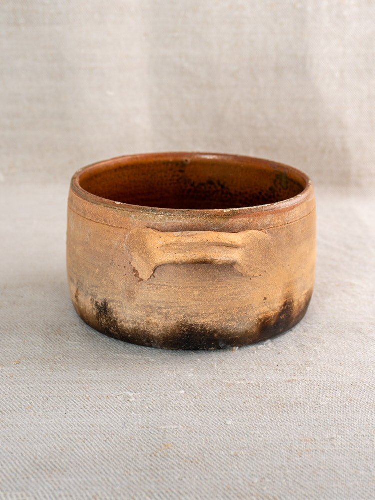 Antique French terracotta hearth cooking pot with bone-shaped motif on textured beige fabric background.
