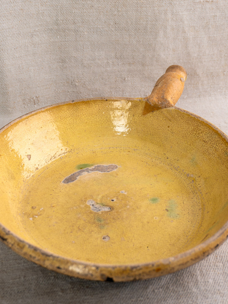 Antique French Aubagne terracotta poêlon with yellow glaze, worn and cracked, on beige textured fabric.