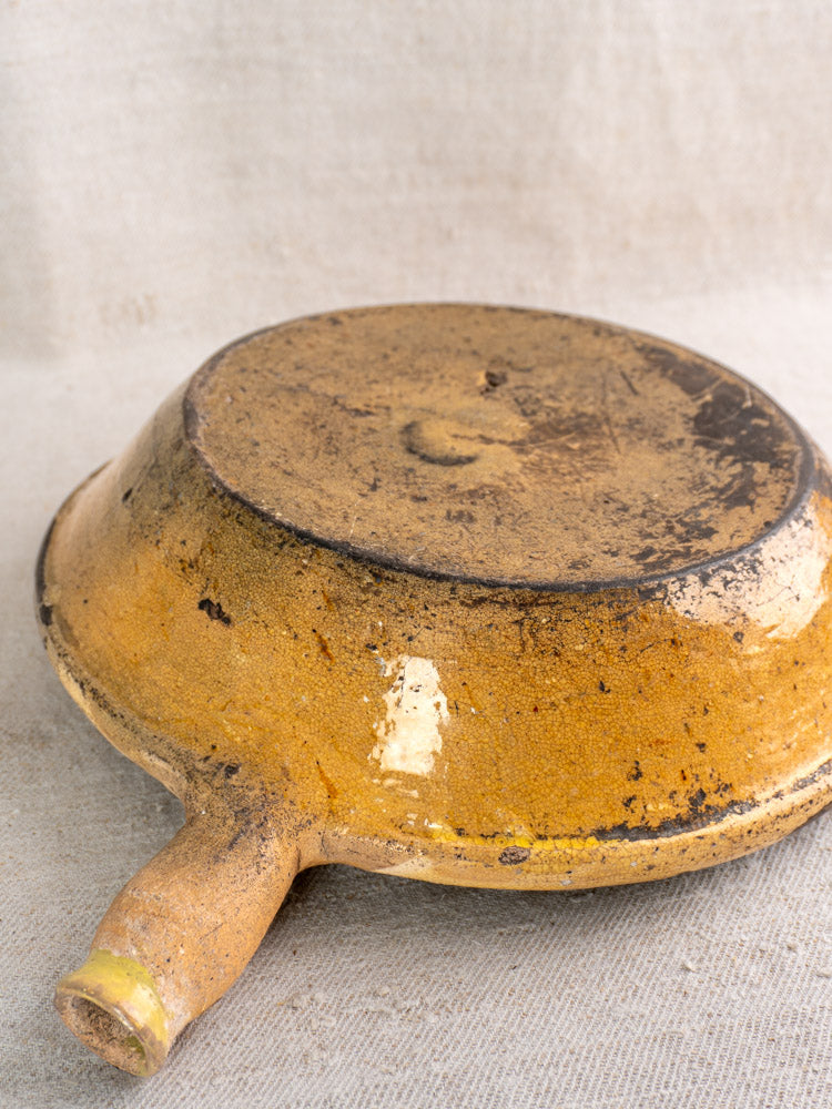 Antique French terracotta poêlon with yellow glaze, late 18th century, inverted on textured beige fabric, showing weathered patina.
