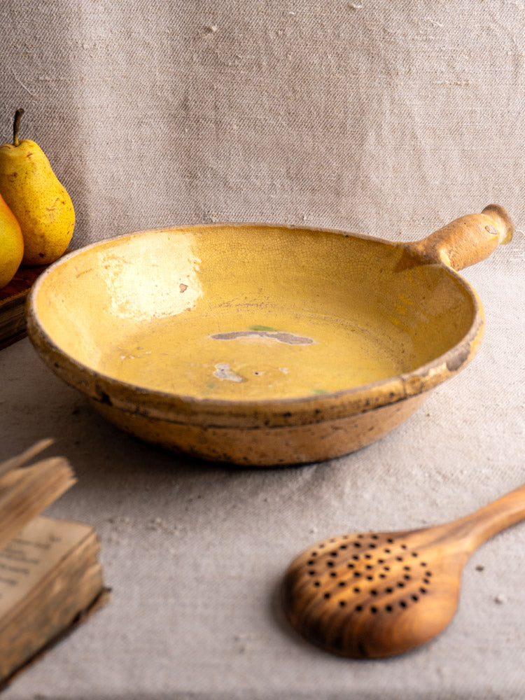 Antique French poêlon from Aubagne, late 18th c., yellow glaze, on beige cloth with wooden spoon and pears.