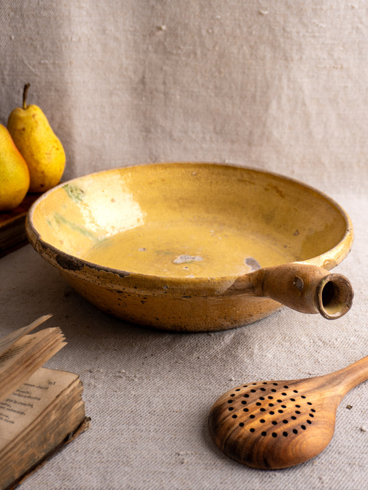 Antique French poêlon from Aubagne, late 18th c., yellow glaze, with wooden slotted spoon and pears on textured surface.