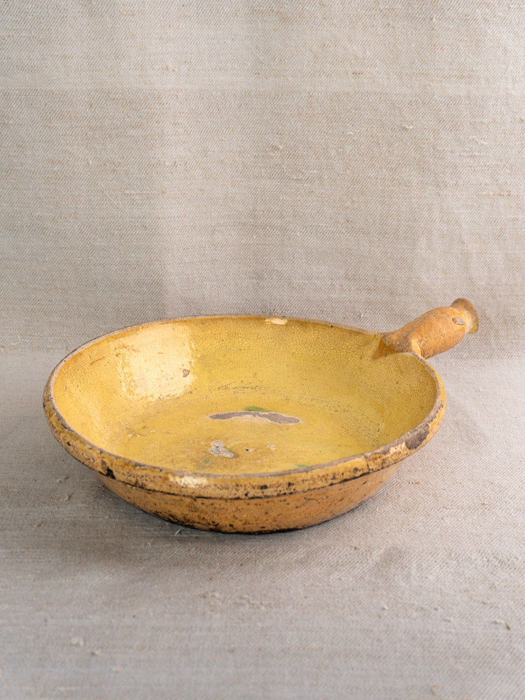 Antique French terracotta poêlon from Aubagne with yellow glaze and small handle on beige textured fabric.