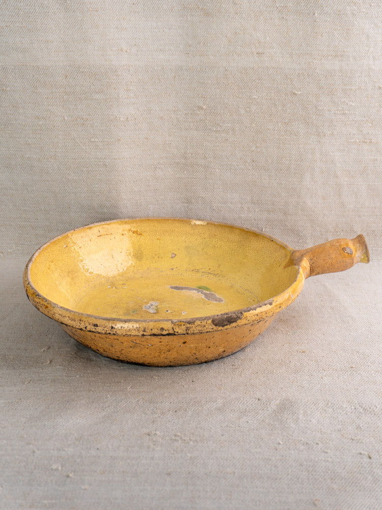 Antique French yellow ceramic poêlon with small handle, visible wear and patina, on beige textured fabric.