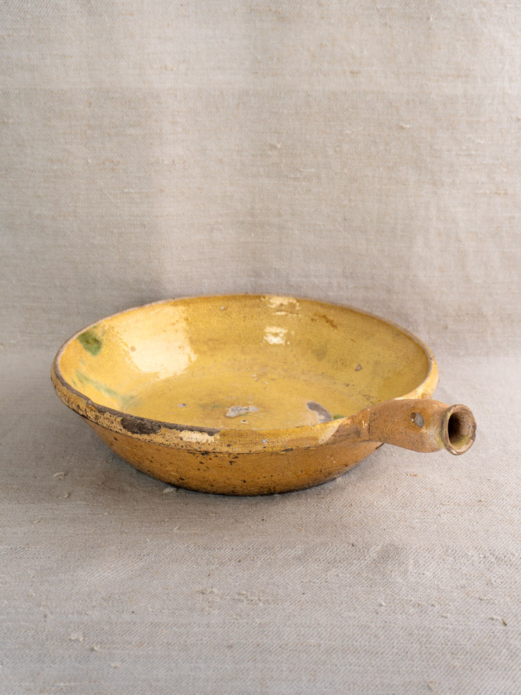 Antique French poêlon from Aubagne in yellow-glazed terracotta with spout handle and aged patina on a textured surface.
