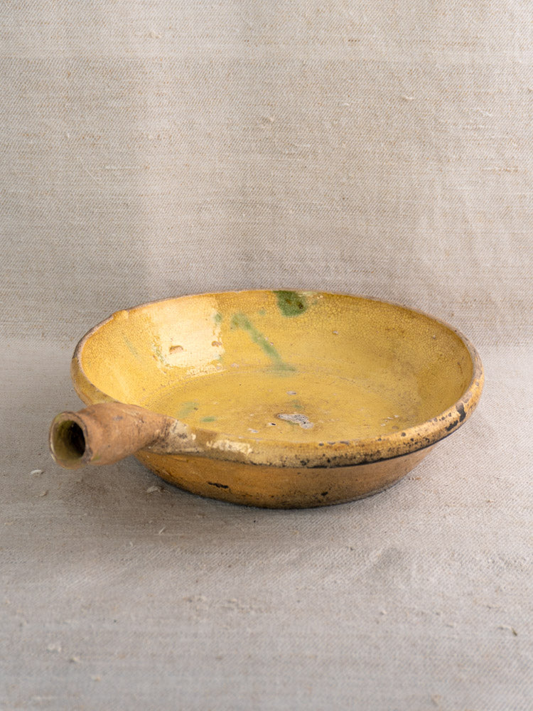 Antique French poêlon from Aubagne with yellow-glazed terracotta, spout, wear, and green spots on neutral textured fabric.