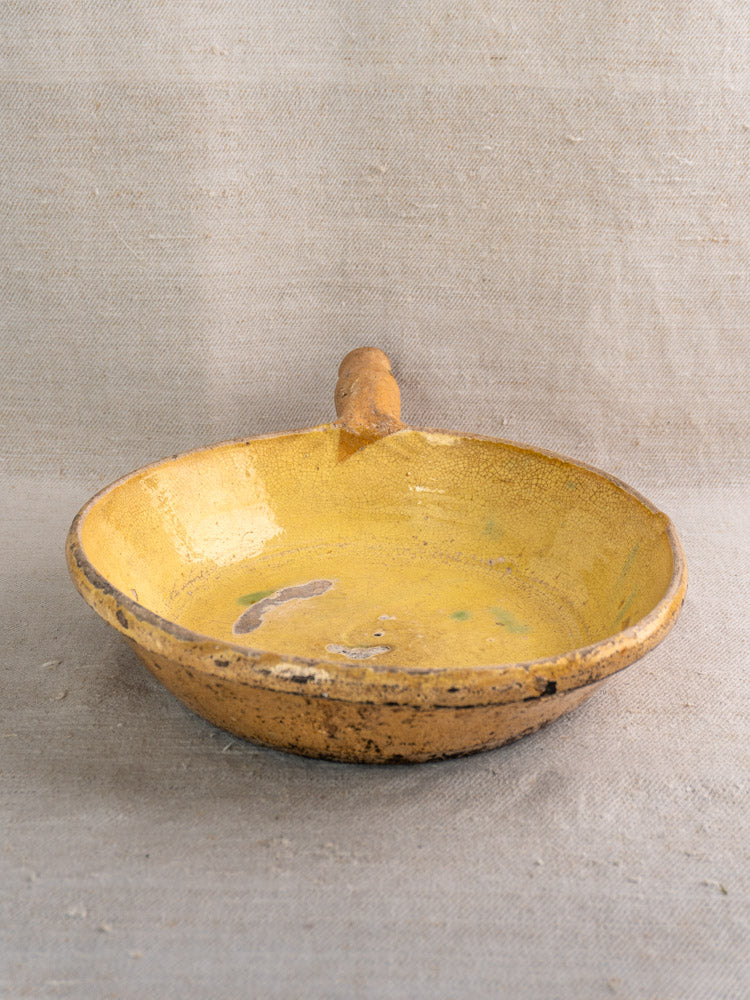 Rustic antique French poêlon, shallow yellow ceramic bowl with handle and chipped edges, on textured beige fabric.