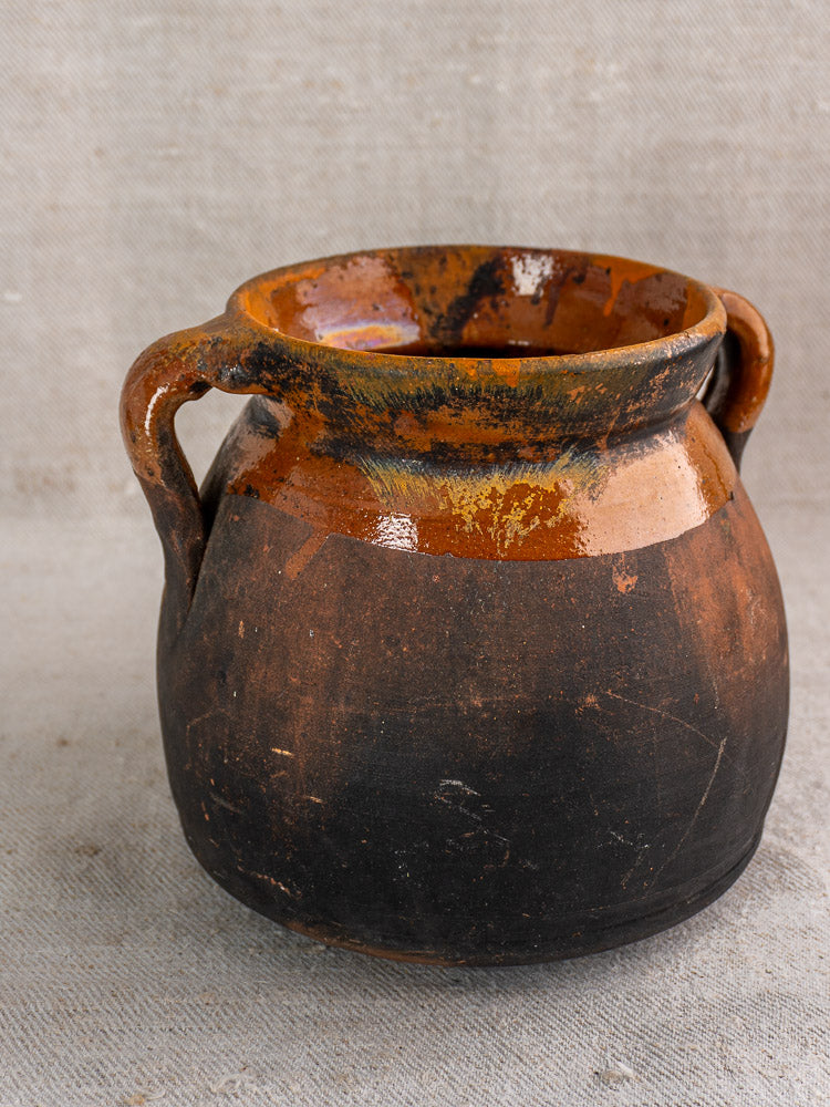 Late 19th-century French confit pot with two handles and brown-black glaze on textured beige fabric.