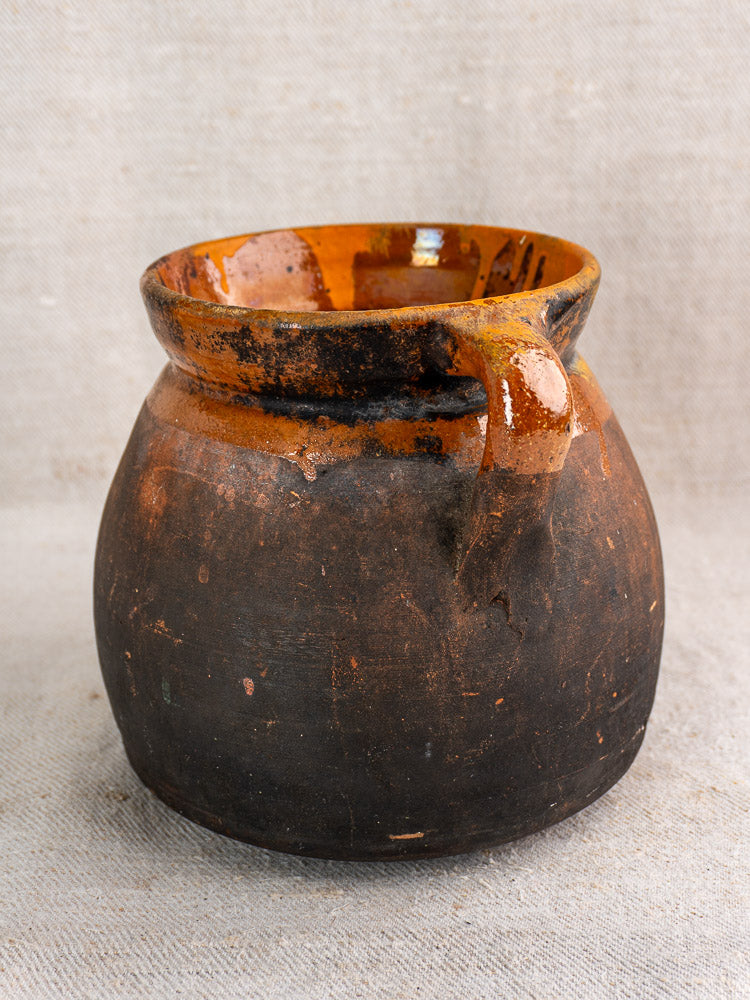 Antique French confit pot with round shape, single handle, orange-brown glazed rim on neutral textured fabric.