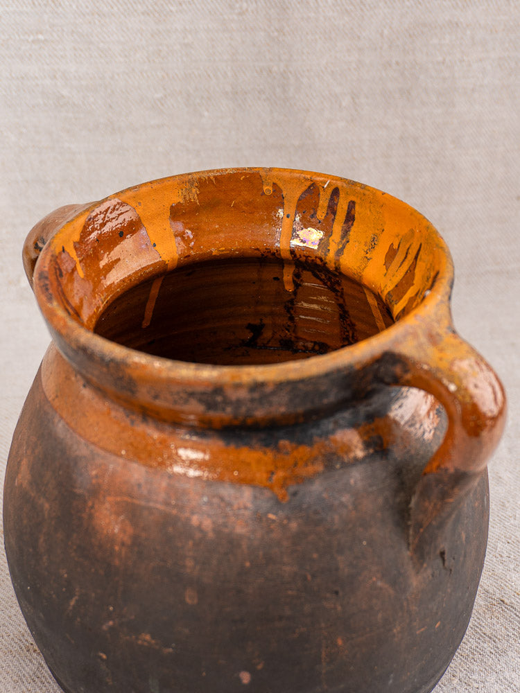 Antique French earthenware confit pot with dual handles, glossy brown rim, matte dark body on neutral fabric.