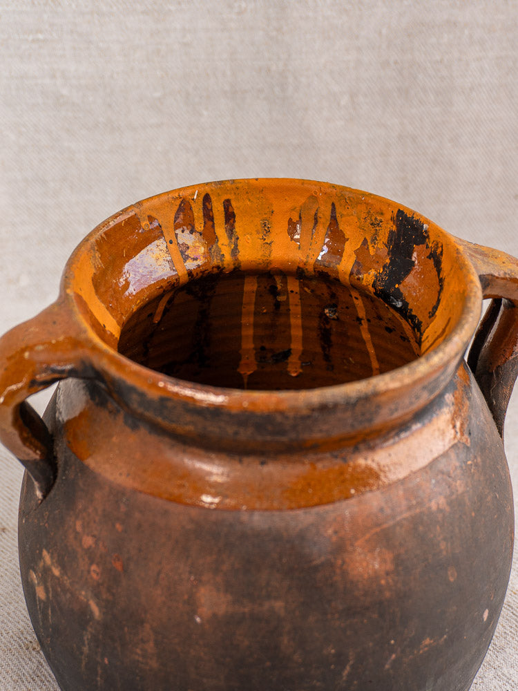 Antique French earthenware confit pot with side handles and glossy orange interior, against a textured beige background.