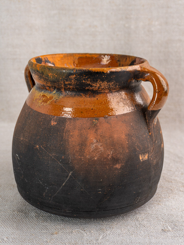 Antique French confit pot, earthenware with dark lower and orange-brown upper halves, on textured light gray fabric.
