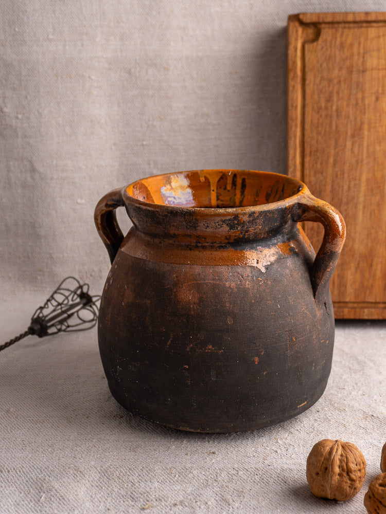 Antique French earthenware confit pot with dark base and brown rim, styled with whisk, board, walnuts on textured fabric.