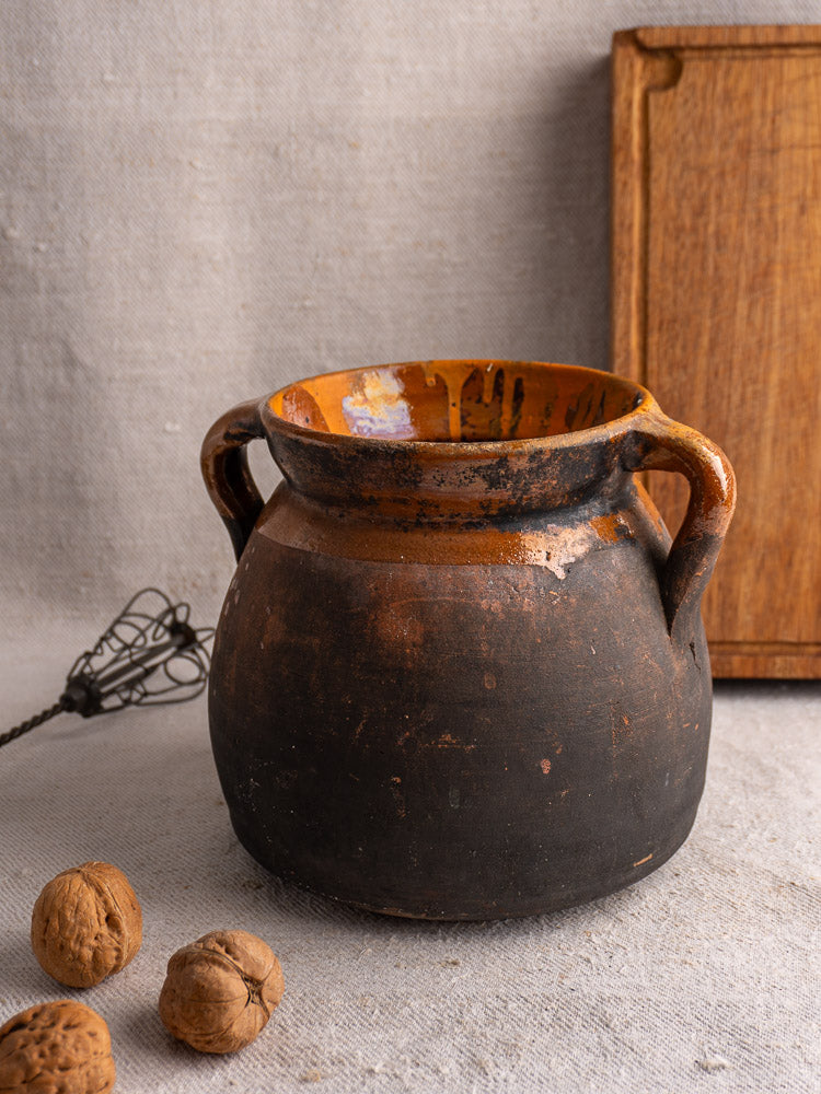 Antique French confit pot, late 19th-century earthenware with dark brown and orange glaze, on beige textured fabric.