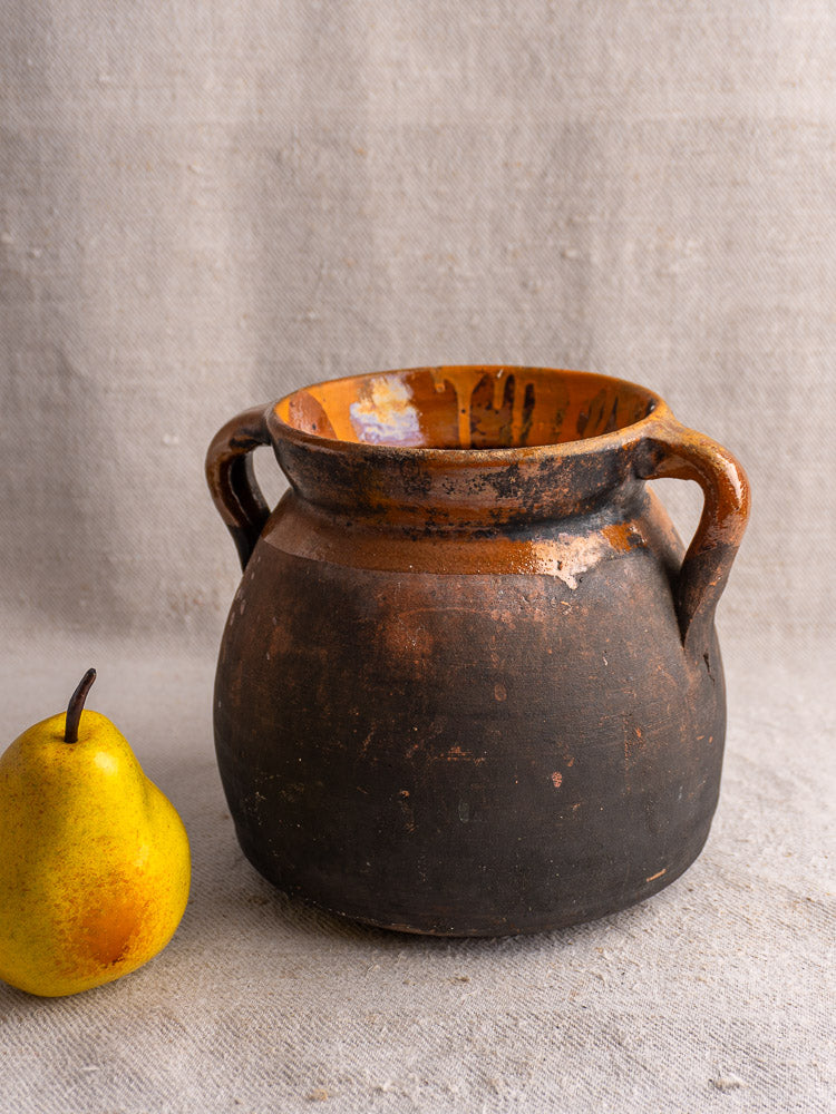 Antique French earthenware confit pot with two handles on beige fabric, paired with a yellow pear.