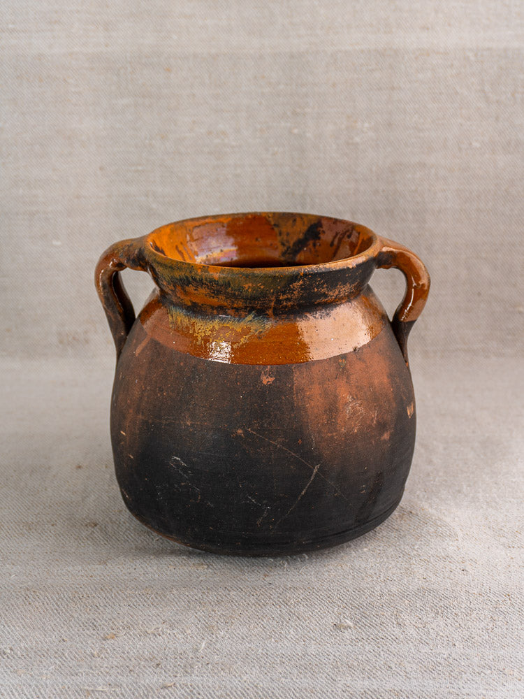 Antique French confit pot, late 19th-century earthenware with dark brown and orange glaze, on beige textured fabric.
