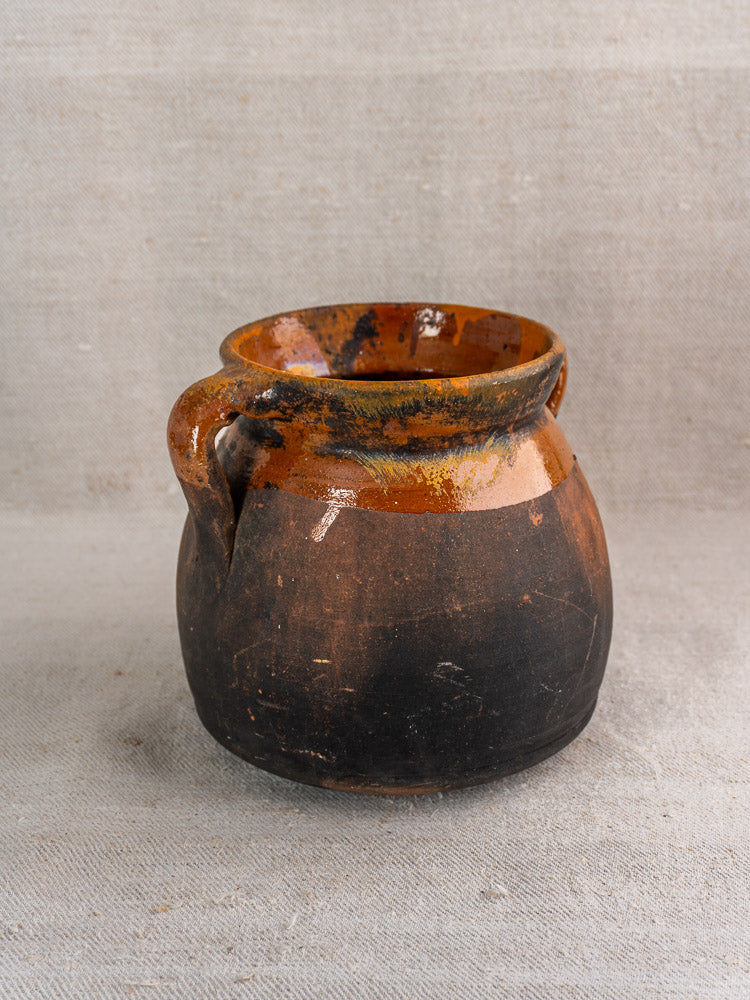 Antique French earthenware confit pot in rustic brown and black, single handle, on textured light gray fabric.