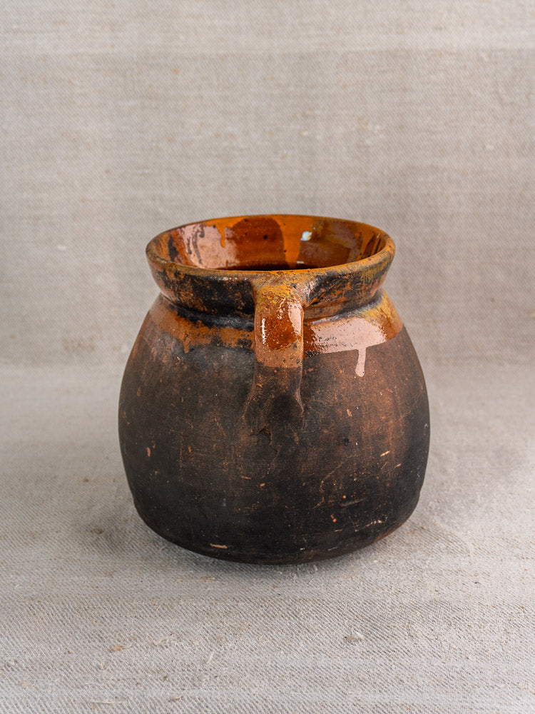 Antique French earthenware confit pot with rustic round form and dark brown finish on beige textured fabric.