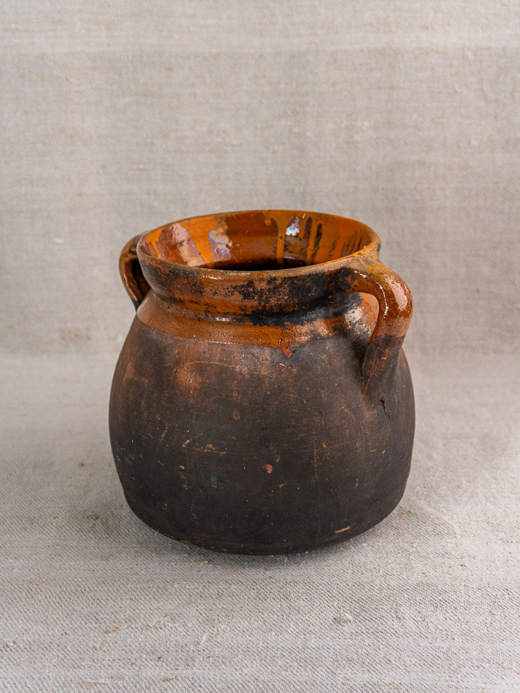 Late 19th century French earthenware confit pot with dark brown exterior, two handles, on neutral fabric background.