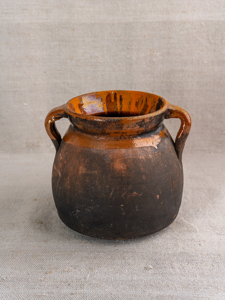 Antique French confit pot in brown earthenware with orange-glazed interior, two handles on textured gray fabric.