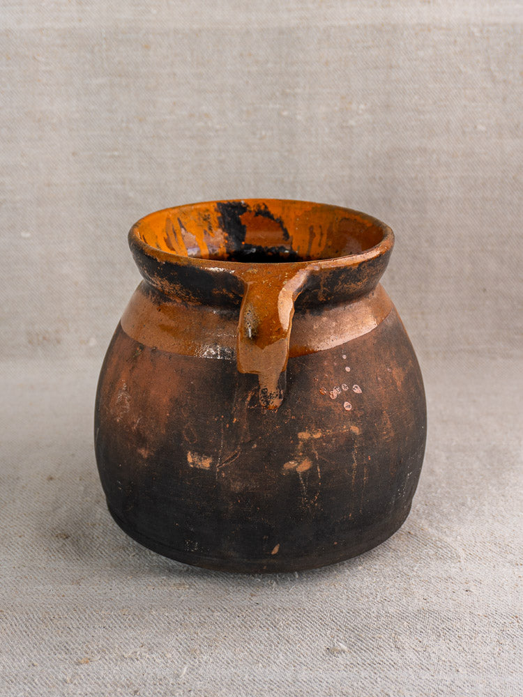 Antique French earthenware confit pot with wide mouth and single handle in brown and orange, on beige textured fabric.