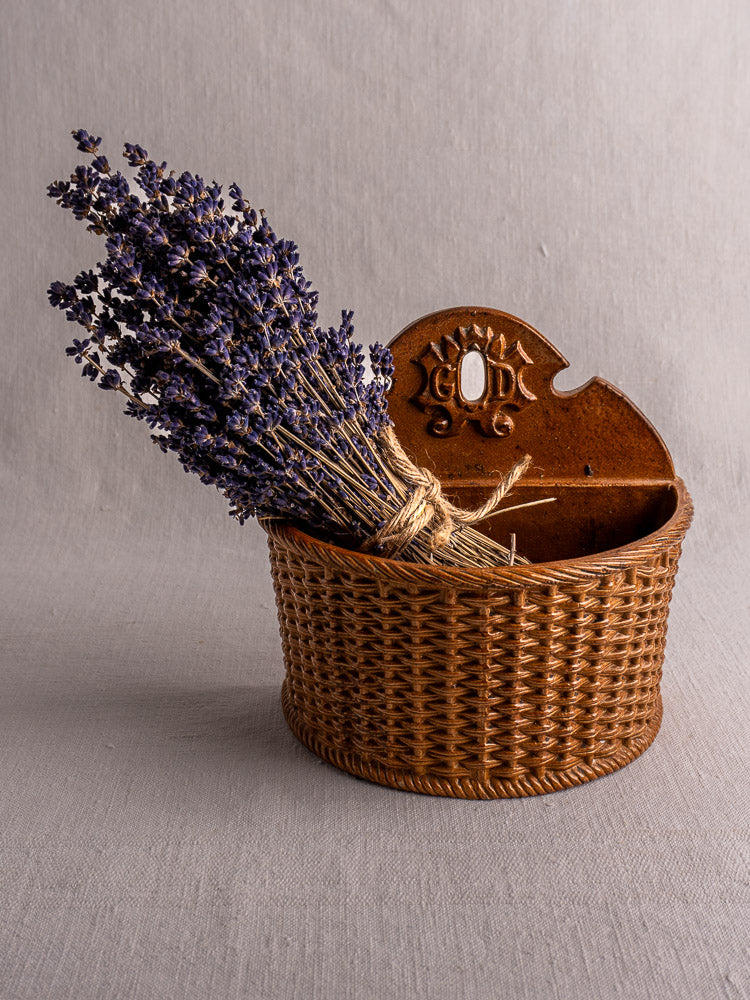 Antique French salt-glazed stoneware salt box with dried lavender in wicker basket, on neutral fabric background.
